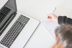 Person writing in a notebook next to an open laptop on a white desk.